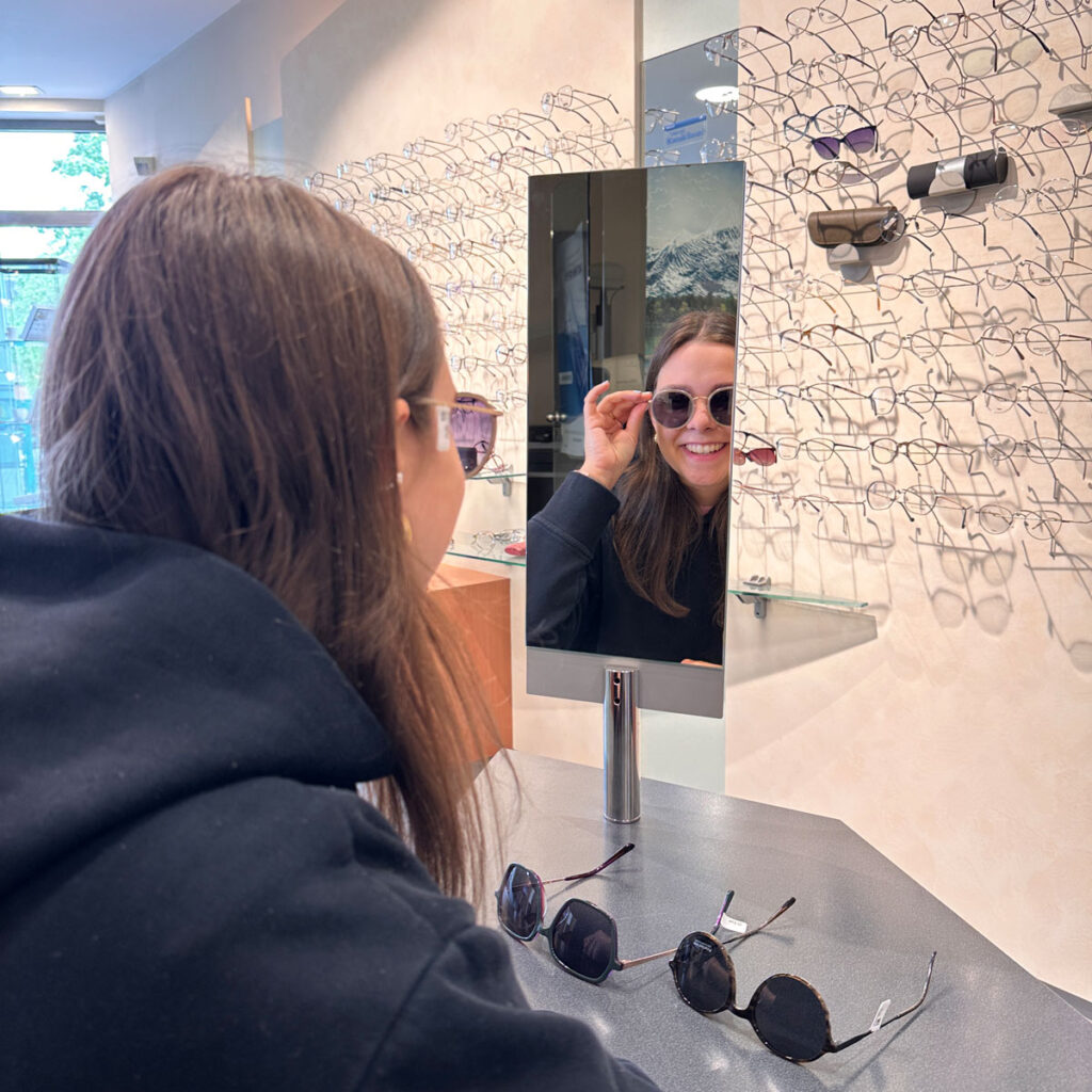 Eine Frau lächelt im Spiegel, probiert Sonnenbrille, trägt schwarzen Pullover in Brillengeschäft.