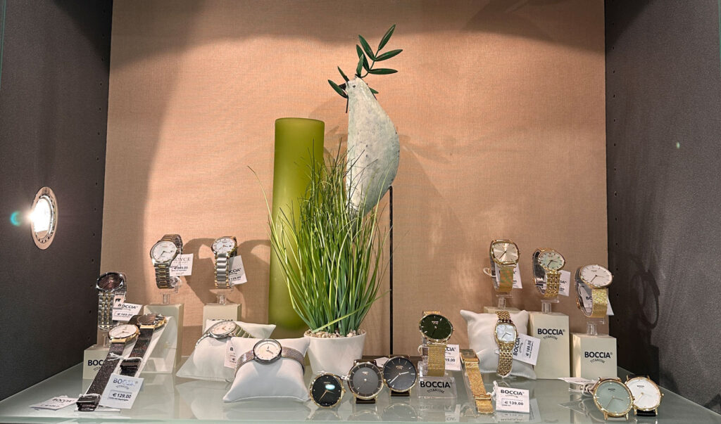 Ein Schaufenster mit diversen Armbanduhren, grünen Dekorvasen und Gras.