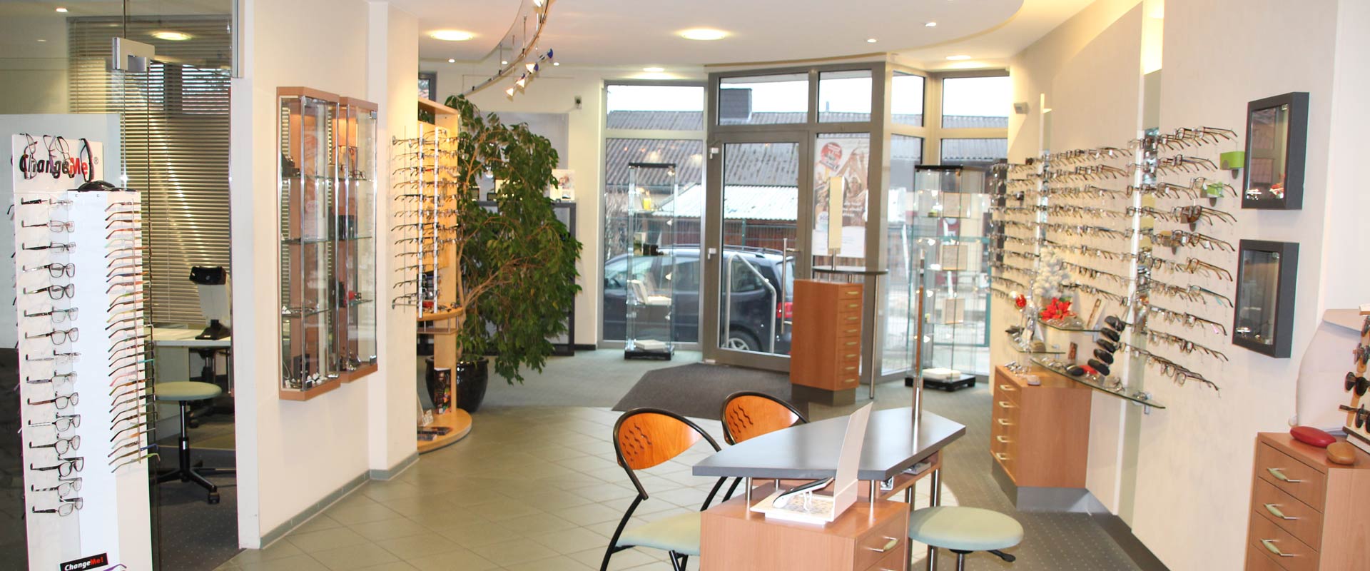 Ein optisches Geschäft mit Brillenregalen, Pflanzen und einem Tisch auf grauem Fliesenboden.